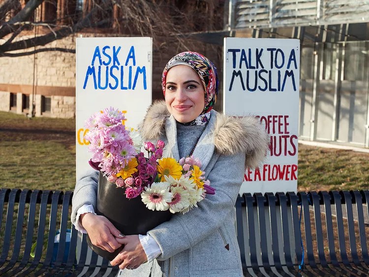A smiling woman in a hijab holds a pot of colorful flowers in front of two signs that say 'Ask a Muslim' and 'Talk to a Muslim' in a public space.